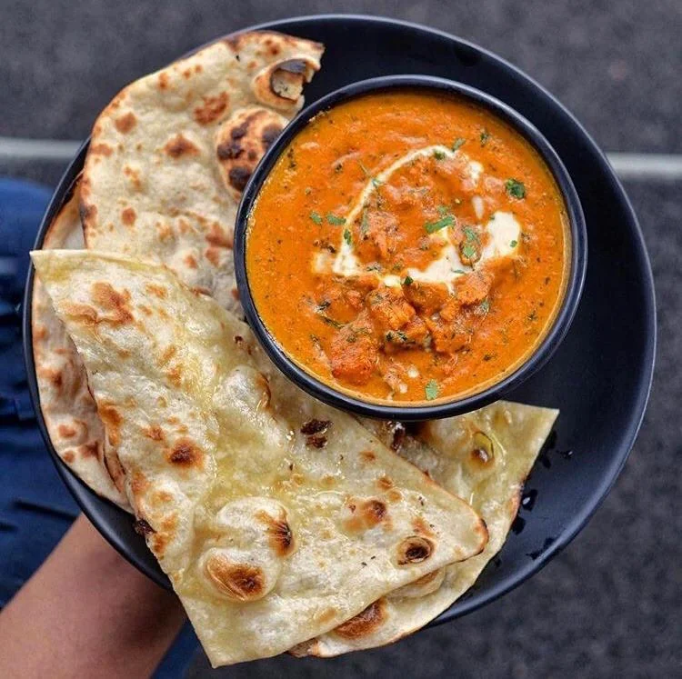 A bowl of creamy butter chicken drizzled with a white sauce and garnished with green herbs next to a fluffy piece of naan.Image from https://www.reddit.com/r/food/comments/d1k6i0/i_ate_butter_chicken_with_naan/ 