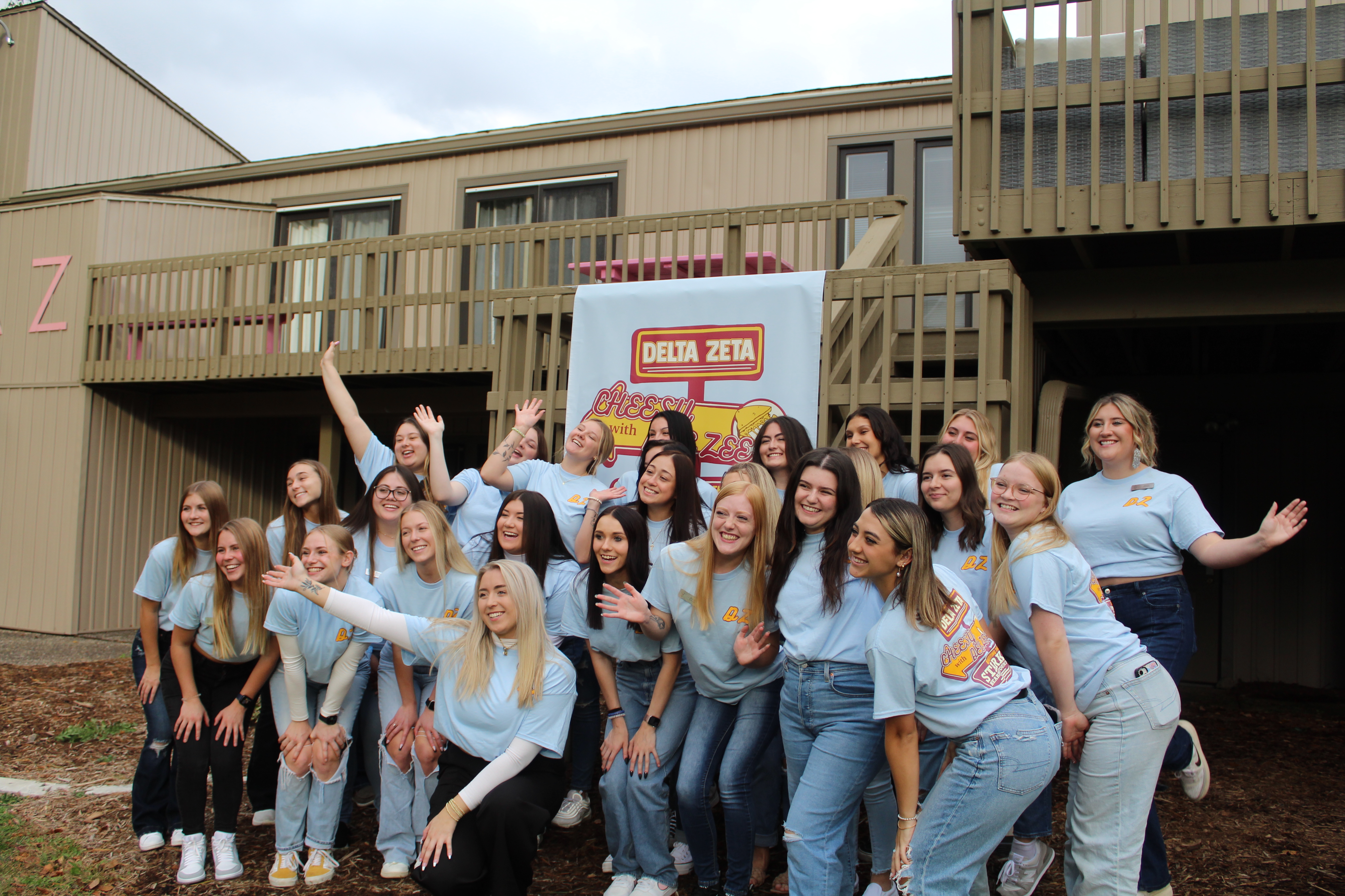 Group of women from the Delta Zeta sorority posing in front of their sorority house. They are wearing matching t-shirts, and they are in front of a banner that promotes their event “Grilled Cheesy with the Dee Zees”. The women are smiling and laughing together. 