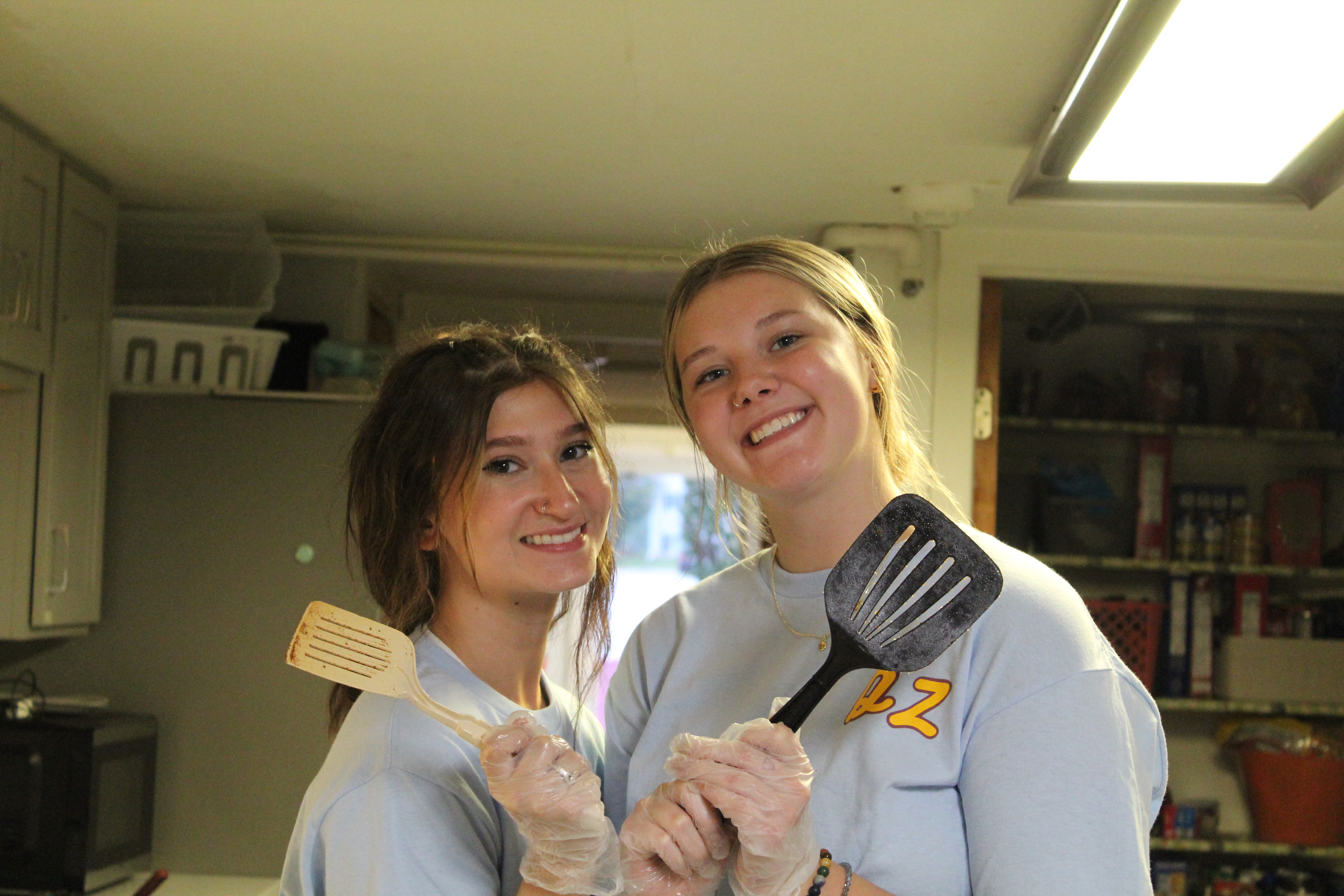 Two women from the Delta Zeta women holding spatulas while smiling in their sorority’s kitchen. 