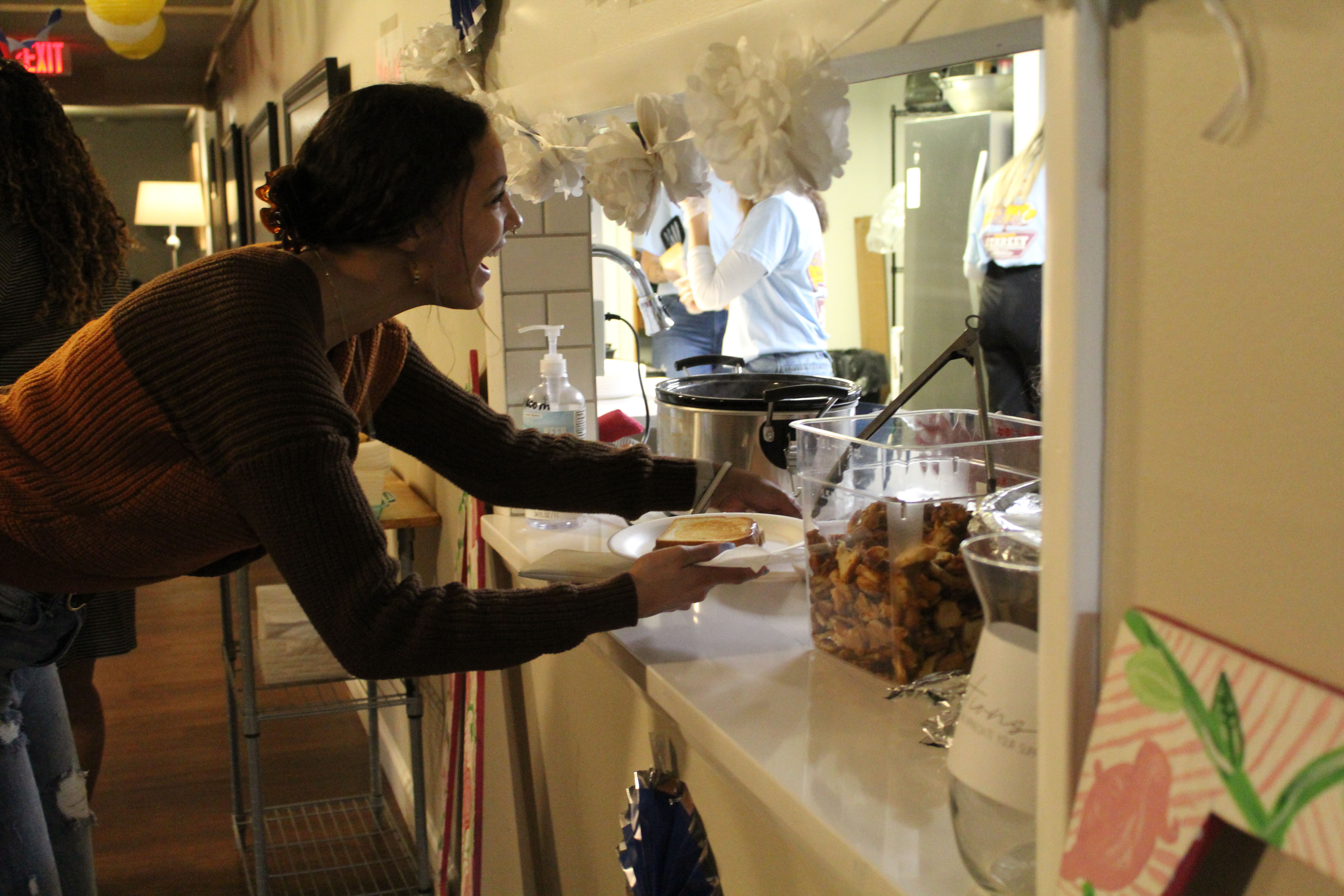 A woman standing in the serving window at the Delta Zeta sorority house. She is holding a grilled cheese and smiling at the women who are standing behind the counter. 