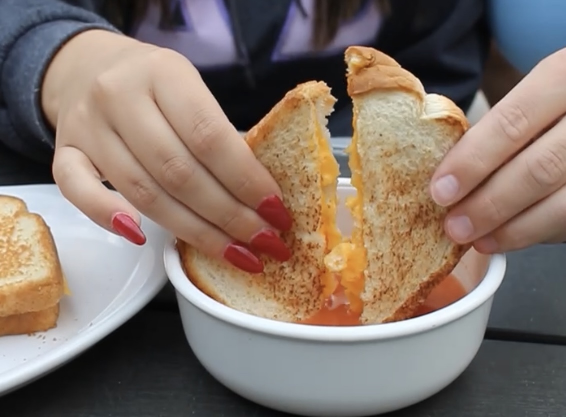 Two hands holding a grilled cheese and dipping it in tomato soup.