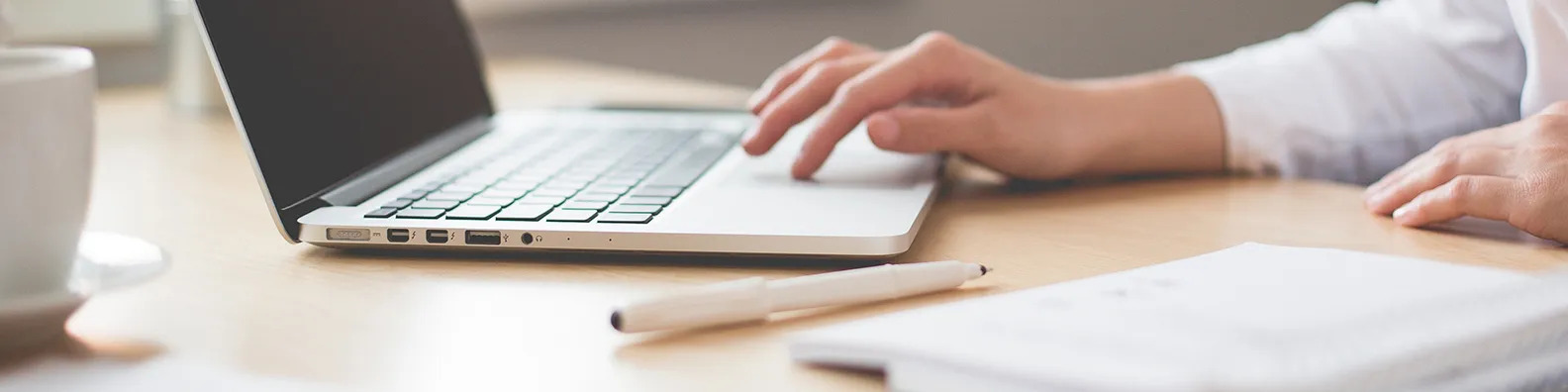 A header image with a open laptop in frame with a pen in the forefront as it appears a woman is working on typing near it. There is also a white coffee mug to the left of the frame.